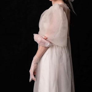 May include: A white wedding dress with sheer, flowing sleeves and a matching veil. The dress features a simple, elegant design, complemented by delicate lace gloves. The overall aesthetic is ethereal and romantic, perfect for bridal wear.