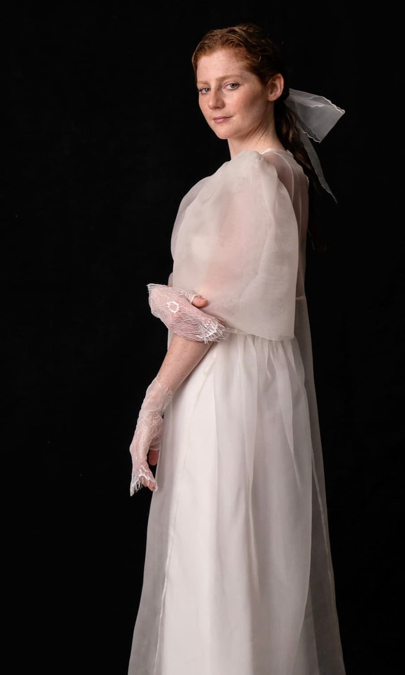 May include: A white wedding dress with sheer, flowing sleeves and a matching veil. The dress features a simple, elegant design, complemented by delicate lace gloves. The overall aesthetic is ethereal and romantic, perfect for bridal wear.