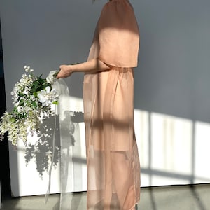 May include: A sheer, peach-colored dress with short sleeves and a gathered waist. The dress is worn with beige block-heeled sandals. A bouquet of white flowers with a white ribbon is held in the hand.