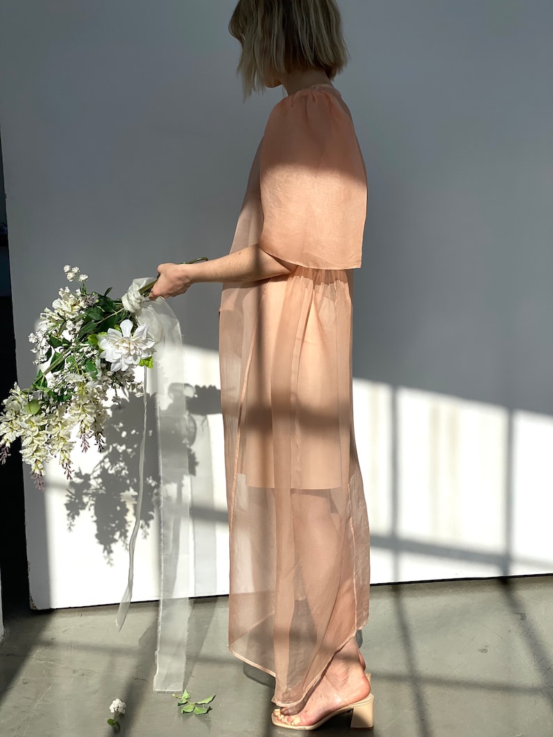 May include: A sheer, peach-colored dress with short sleeves and a gathered waist. The dress is worn with beige block-heeled sandals. A bouquet of white flowers with a white ribbon is held in the hand.