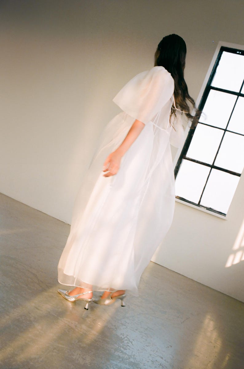 May include: A white, flowing dress with sheer, puffy sleeves and a long skirt. The dress is paired with silver heels. The image is taken in a room with a window, and the lighting creates shadows on the floor.