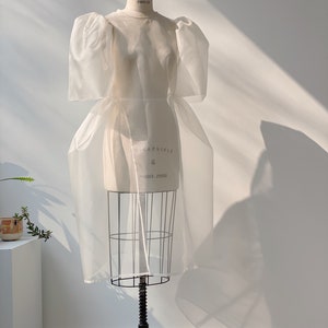 May include: A white, sheer dress with puffy sleeves is displayed on a dress form. The dress form is on a black, wheeled stand. The dress has a fitted bodice and a flowing skirt. The dress form has the text "COLLAPSIBLE" and "MODEL 2000".
