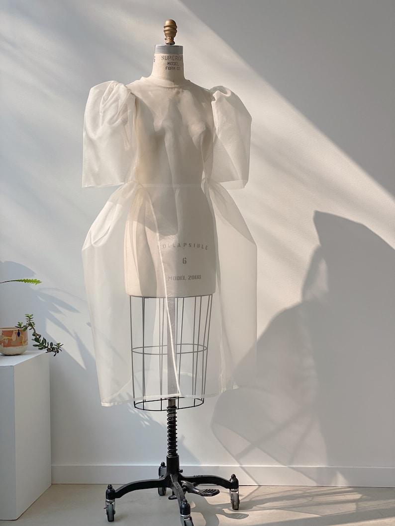 May include: A white, sheer dress with puffy sleeves is displayed on a dress form. The dress form is on a black, wheeled stand. The dress has a fitted bodice and a flowing skirt. The dress form has the text "COLLAPSIBLE" and "MODEL 2000".