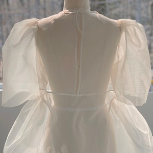 May include: A white, sheer dress on a dress form. The dress features puffed sleeves and a flowing skirt. The fabric appears lightweight and delicate, with a subtle sheen. The dress is designed with a high neckline and a fitted waist.