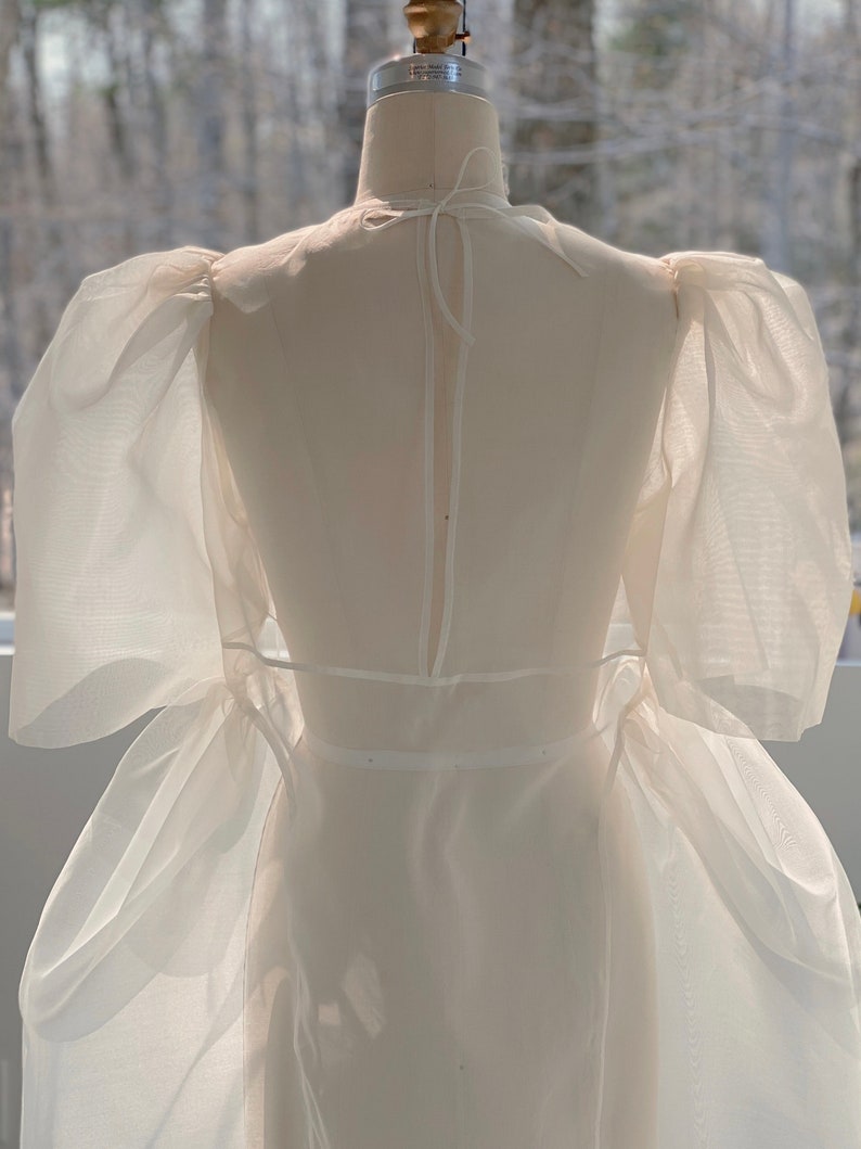May include: A white, sheer dress on a dress form. The dress features puffed sleeves and a flowing skirt. The fabric appears lightweight and delicate, with a subtle sheen. The dress is designed with a high neckline and a fitted waist.