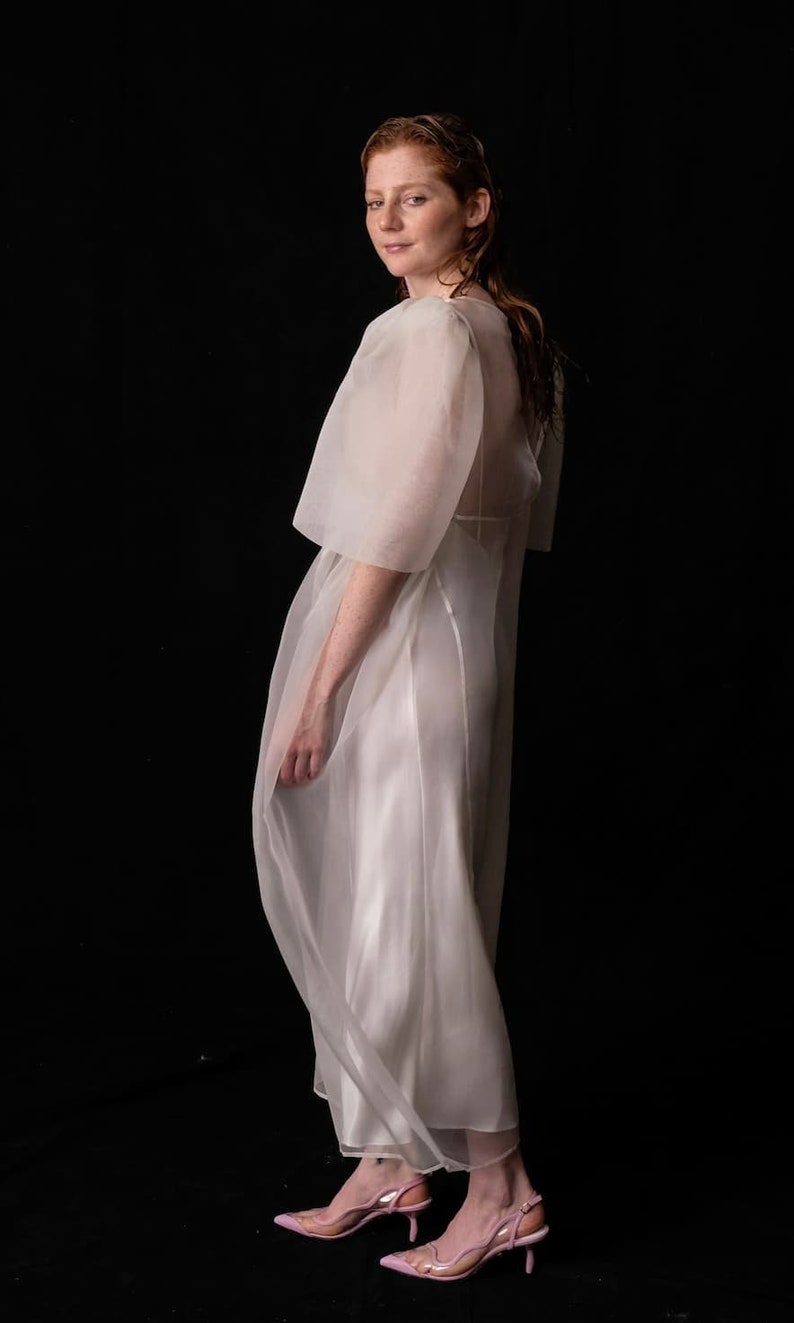 May include: A sheer white dress with short sleeves and a flowing skirt. The dress is paired with pink heels with clear straps. The background is solid black, creating a contrast with the light-colored dress.