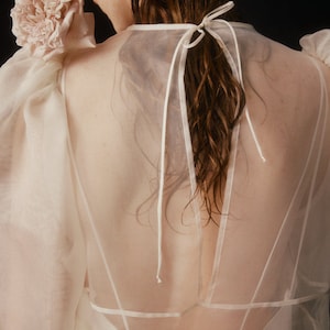 May include: A sheer, cream-colored top with a delicate, flowing design. The top features puffy sleeves and a low back, with a bow detail at the neck. A large, light pink flower adorns the shoulder, adding a touch of elegance.