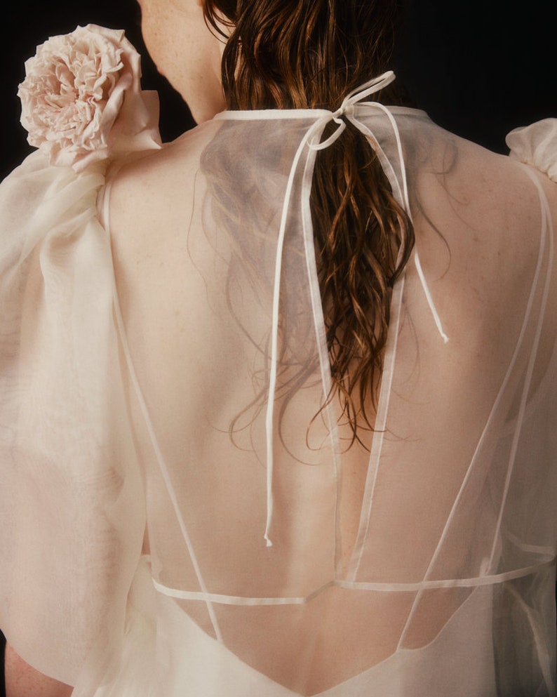 May include: A sheer, cream-colored top with a delicate, flowing design. The top features puffy sleeves and a low back, with a bow detail at the neck. A large, light pink flower adorns the shoulder, adding a touch of elegance.