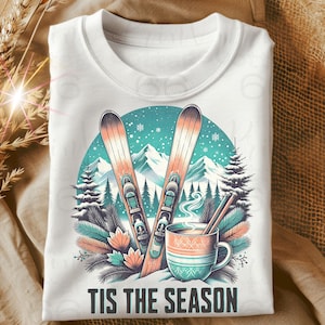 May include: White crew neck sweatshirt with a winter-themed graphic. The design features skis, a mug of hot beverage, and the text "TIS THE SEASON" in a winter landscape setting. The graphic is in shades of teal, orange, and white.