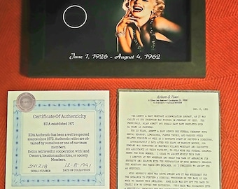 May include: A black framed display containing a hair strand of Marilyn Monroe, featuring her portrait and the text Authentic hair strand of Marilyn Monroe, June 1, 1926 - August 4, 1962, alongside a Certificate of Authenticity and a letter.
