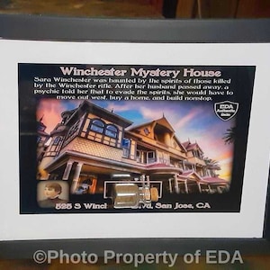 May include: A framed photo of the Winchester Mystery House in San Jose, California. The photo shows the exterior of the house, which is a large, Victorian-style mansion with many windows and turrets. The text on the photo reads "Winchester Mystery House" and "525 S Winch vd, San Jose, CA". The photo is framed in a black frame and has a white mat.
