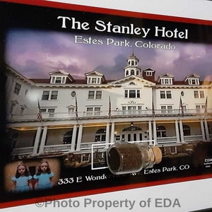 May include: A framed photograph of The Stanley Hotel in Estes Park, Colorado. The hotel is a white building with a large porch and a tower. The photo is framed in a black frame with a red mat. The text "The Stanley Hotel" and "Estes Park, Colorado" is printed on the mat. The text "Photo Property of EDA" is printed on the bottom of the frame.