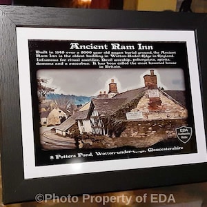 May include: A framed print of the Ancient Ram Inn, a historic building in Wotton-Under-Edge, England. The black frame surrounds a picture with text describing the inn's history, including its reputation as a haunted location. The print includes the text "Ancient Ram Inn".