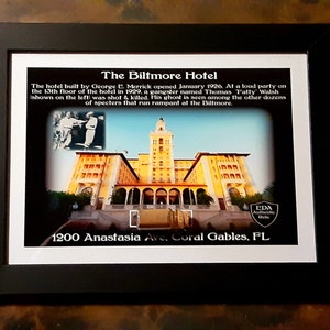 May include: Framed print of The Biltmore Hotel in Coral Gables, FL. The image features the hotel's exterior, a vintage photo of two men, and text describing the hotel's history, including a shooting and ghostly sightings. The frame is black.