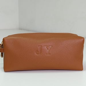 Könnte beinhalten: Eine rechteckige, braune Kulturtasche aus Leder mit Reißverschluss und kleinem Griff an der Seite. Die Initialen "JY" sind auf der Vorderseite eingeprägt. Die Tasche ist aus strukturiertem Leder.