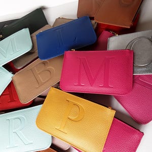 Puede incluir: Una colección de bolsos con cremallera de cuero de colores, cada uno grabado con una sola letra. Los bolsos son de varios tonos, incluyendo azul, rosa, amarillo y beige. Las letras son en negrita, sin serifa.