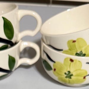 May include: Four white ceramic teacups stacked in pairs. Each cup features a yellow floral design with green leaves and black stems. The cups have curved handles and a slightly angled rim.
