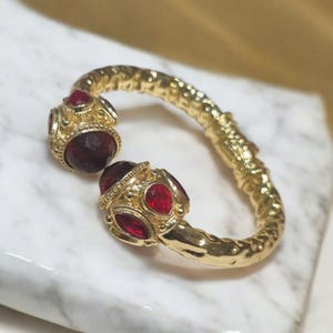 Könnte beinhalten: Ein goldfarbenes Armreif-Armband mit strukturiertem Design und zwei roten Edelstein-Akzenten.