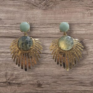 May include: A pair of gold-toned earrings with a light green gemstone at the top and a larger, round, patterned gemstone at the center. The earrings have a sunburst design with a textured, antique finish. The background is a wooden surface.