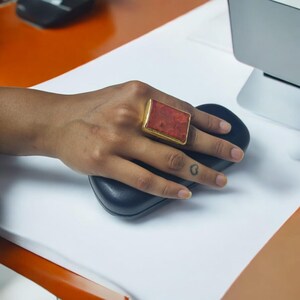 May include: A gold ring with a large, rectangular red stone set in the center. The ring is worn on the index finger of a hand.