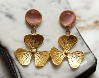 May include: A pair of gold-colored earrings with a floral design. Each earring features a round, light pink stone at the top, connected to a three-petal flower. The earrings are displayed on a white and gray marble surface.