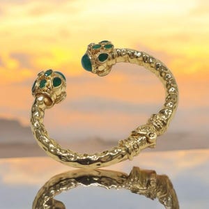 May include: A gold bangle bracelet with two green gemstone accents. The bracelet is textured and has a hammered finish.