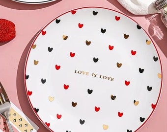 May include: White ceramic dinner plate with a red rim. The plate is decorated with a pattern of red, black, and gold hearts. The text "LOVE IS LOVE" is printed in gold in the centre of the plate.