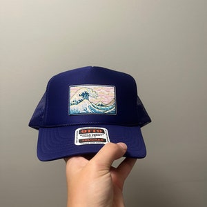May include: A navy blue trucker hat with a mesh back. The front of the hat features a patch with a colorful image of a wave. The patch also has the text "OTTO WORLD FAMOUS FOAM FRONT SNAPBACK TRUCKER" on it.