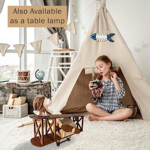 May include: A wooden airplane-shaped table lamp with a star design on the side. The lamp is brown with a light-colored interior and propeller. The text "Also Available as a table lamp" is in the top left corner.