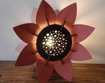 Puede incluir: Lámpara decorativa con forma de flor, con pétalos rosas y rojos superpuestos. El centro negro tiene pequeños agujeros circulares que emiten una luz cálida. Una lámpara decorativa.