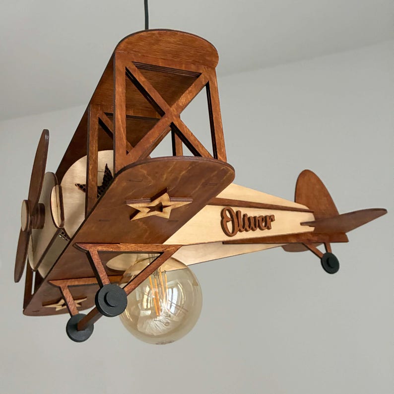 May include: A wooden airplane-shaped pendant light fixture. The fixture is made of light and dark brown wood, with a propeller, wings, and a star-shaped cutout. The word "Oliver" is written on the side. A large, round light bulb hangs below.