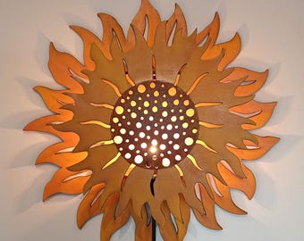 Puede incluir: Una lámpara de pared de madera con forma de girasol. La luz tiene un diseño detallado con un centro de pequeños agujeros y un patrón de pétalos circundantes. La lámpara está pintada en tonos naranja y marrón, con un cable de alimentación negro.