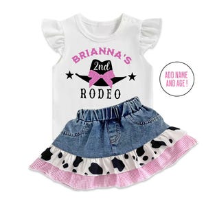 May include: White t-shirt with pink glitter text that says "Brianna's 2nd Rodeo" with a black cowboy hat and pink bow. The shirt is paired with a denim skirt with a white and black cow print ruffle.