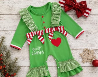 May include: A bright green long-sleeve romper featuring ruffled shoulders and leg cuffs, a red heart patch, and the white text "my 1st Christmas". The romper includes a red and white striped waist tie and a matching red sequined bow headband.