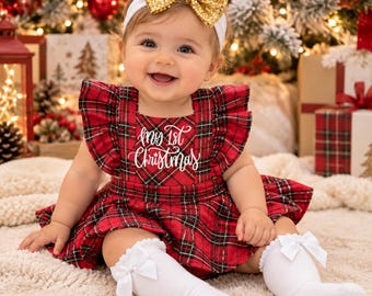 May include: A red and green plaid jumper dress featuring the white embroidered text My 1st Christmas, paired with a white headband topped with a gold sequin bow and white ribbed socks accented with small white bows at the cuffs.