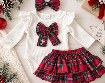 May include: A white ribbed long-sleeve bodysuit with ruffled shoulders, featuring a large red and green plaid bow with a small silver cross charm. Includes a matching red and green plaid skirt and a coordinating plaid headband bow.
