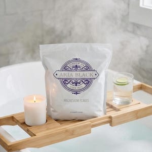 May include: A white bag of Aria Black magnesium flakes sits on a bamboo bath tray with a lit candle and a glass of water with a lime slice. The bag has a circular logo with text.