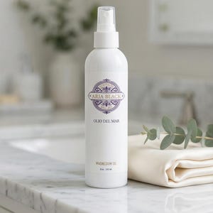 May include: A white spray bottle of Aria Black Magnesium Oil, 8 oz / 250 mL, with a purple and gold label. The bottle is on a marble surface next to a folded cream-colored cloth and sprig of eucalyptus.