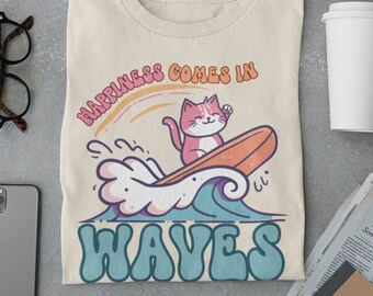 May include: A beige t-shirt with a graphic of a pink cat surfing on a wave. The text "Happiness comes in waves" is printed in a retro font.