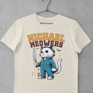 May include: A beige t-shirt with a graphic of a white cat holding a knife. The cat is wearing blue overalls and has a menacing expression. The text "MICHAEL MEOWERS" is printed in yellow above the cat.