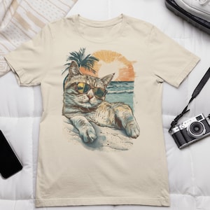 Könnte beinhalten: Ein beiges T-Shirt mit einem grafischen Aufdruck eines Katzen, das eine Sonnenbrille trägt und sich an einem Strand mit Palmen und Sonnenuntergang im Hintergrund entspannt.