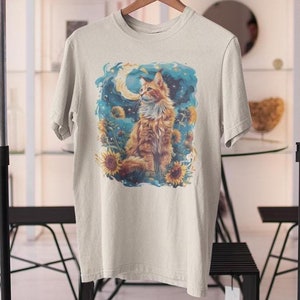 Könnte beinhalten: Ein beiges T-Shirt mit einem bunten Grafikdruck eines Katers, der in einem Feld mit Sonnenblumen unter einem Halbmond sitzt. Der Kater ist orange und weiß mit blauen Augen.