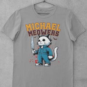May include: A gray t-shirt with a graphic of a white cat holding a knife. The cat is wearing blue overalls and has a menacing expression. The text "MICHAEL MEOWERS" is printed above the cat.