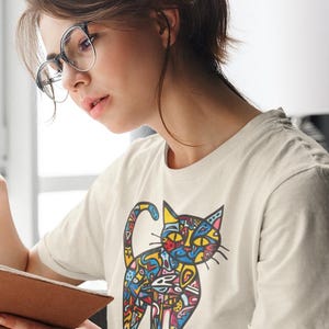 May include: Cream-colored t-shirt featuring a colorful, abstract cat design. The cat illustration uses a variety of colors and patterns. The person in the image is wearing glasses and is holding a book.