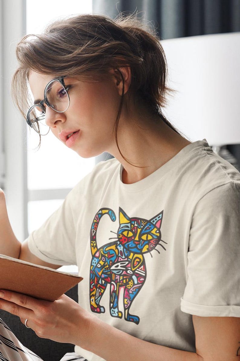 May include: Cream-colored t-shirt featuring a colorful, abstract cat design. The cat illustration uses a variety of colors and patterns. The person in the image is wearing glasses and is holding a book.