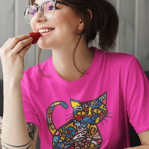 May include: A bright pink t-shirt featuring a colorful, abstract cat design. The cat illustration uses a variety of colors including blue, yellow, and red. The person in the image is wearing glasses and eating a strawberry.