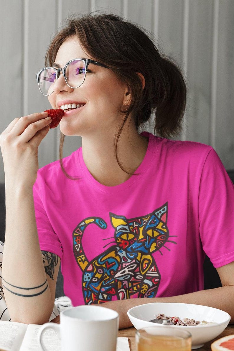 May include: A bright pink t-shirt featuring a colorful, abstract cat design. The cat illustration uses a variety of colors including blue, yellow, and red. The person in the image is wearing glasses and eating a strawberry.
