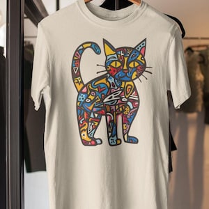 May include: A cream-colored t-shirt featuring a colorful, abstract cat design. The cat illustration is composed of various shapes and patterns in shades of blue, yellow, red, and green. The shirt is hanging on a wooden hanger.