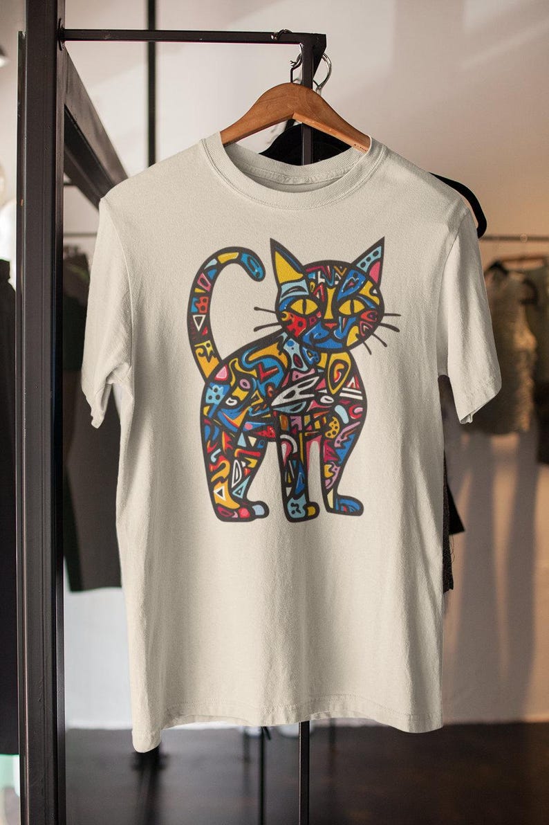 May include: A cream-colored t-shirt featuring a colorful, abstract cat design. The cat illustration is composed of various shapes and patterns in shades of blue, yellow, red, and green. The shirt is hanging on a wooden hanger.