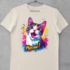 May include: A cream-colored t-shirt featuring a colorful graphic of a cat with the word "Meow!" The cat has blue eyes, and its fur is a mix of colors. The graphic has pink, blue, and yellow paint splatters.